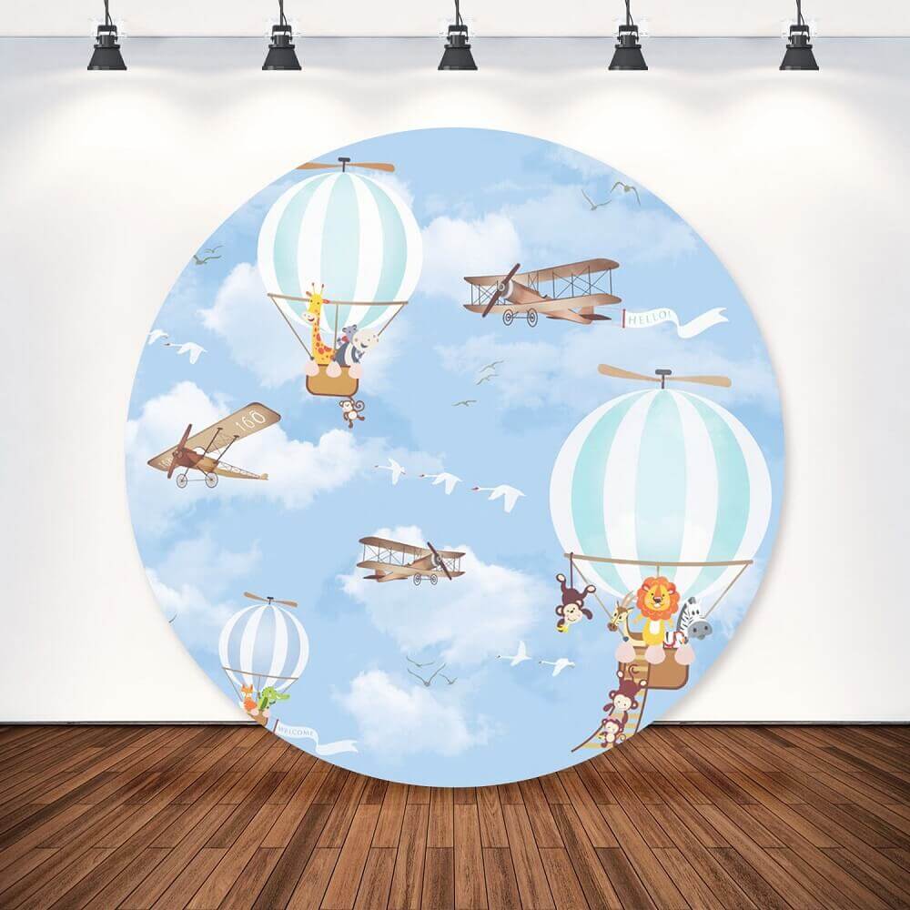 A round backdrop featuring a playful design with hot air balloons, adorable animals, and vintage airplanes in a scenic blue sky with fluffy clouds."