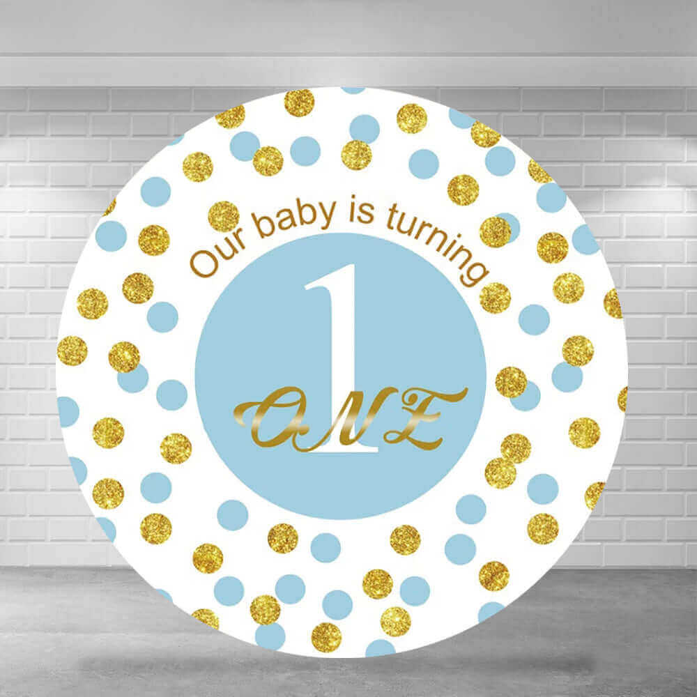 A round baby's first birthday party backdrop with a blue and gold color scheme, featuring the text 'Our baby is turning 1' and the number '1' in a large font, surrounded by glittery gold and pastel blue dots, set against a white brick wall background, ideal for celebrating a baby boy's first birthday.