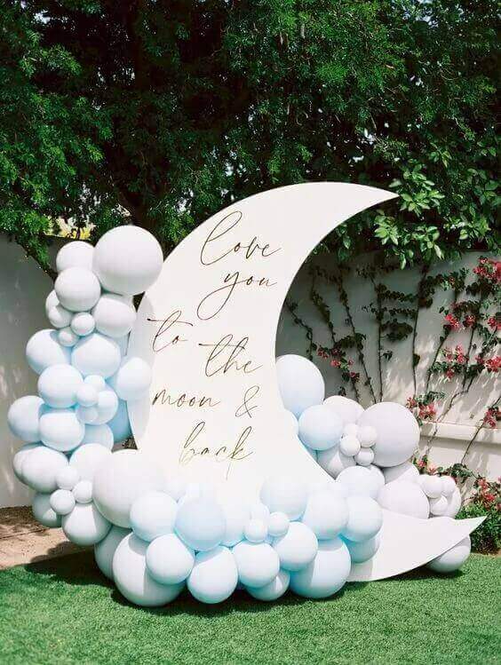 A crescent moon-shaped white backdrop featuring elegant calligraphy that reads "Love you to the moon & back." The setup is decorated with clusters of soft blue and white balloons, placed outdoors against a green lawn with a tree and a flowering vine-covered wall in the background.