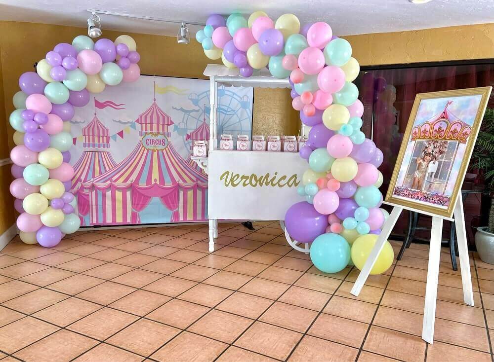 Circus-themed birthday party decor featuring a pastel circus tent backdrop with Ferris wheel, colorful bunting, and a whimsical balloon garland in pink, purple, yellow, and mint green, perfect for carnival-themed celebrations.