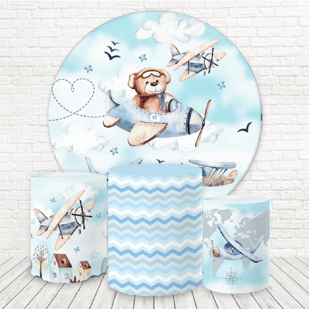 A bear pilot-themed party decor set featuring a circular backdrop with a teddy bear flying an airplane through the clouds, and three decorative cylinder covers with airplane, chevron, and map designs.