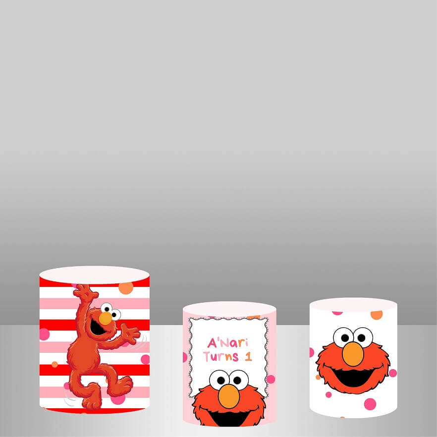 Three cylindrical covers with an Elmo theme displayed against a gray background. The left cylinder features Elmo with outstretched arms on a red and white striped background. The center cylinder reads "A'Nari Turns 1" with Elmo's face below the text on a pink polka-dotted background. The right cylinder displays Elmo's face on a white background with colorful polka dots.