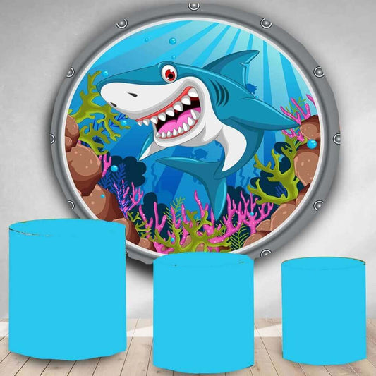A circular cartoon ocean backdrop displaying a cheerful shark surrounded by a colorful coral reef, accompanied by three blue cylinder covers, ideal for kids' parties with an underwater adventure theme