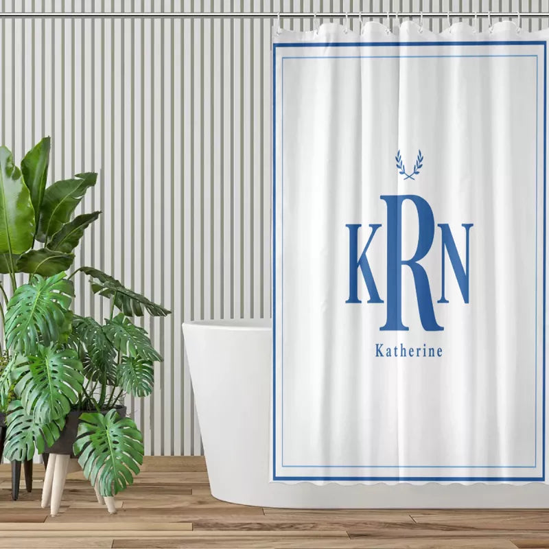 White shower curtain with a blue double-line border, KRN monogram, laurel motif, and Katherine name displayed in a modern bathroom with green plants.