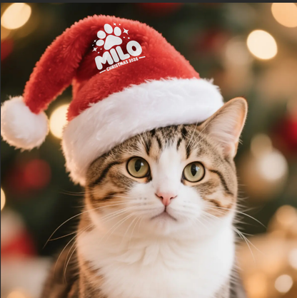Tabby cat wearing a personalized red Santa hat with a white MILO paw print design against a festive Christmas background.