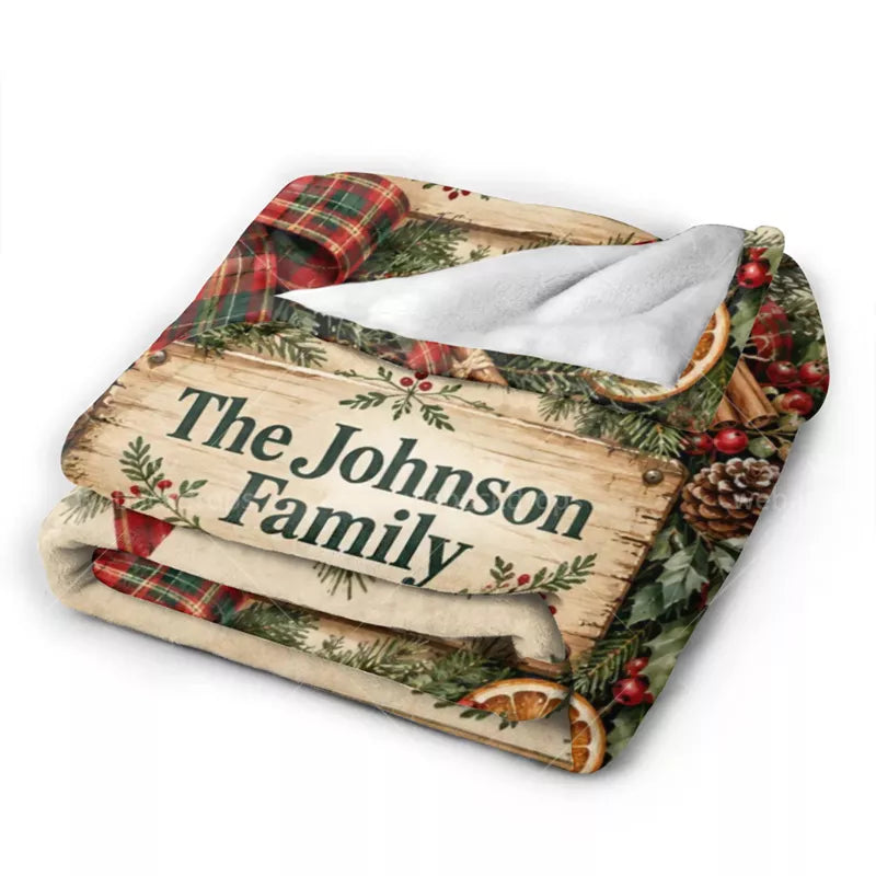 Folded personalized Christmas flannel blanket showing the rustic family name sign, festive plaid artwork, printed greenery, and white reverse side.