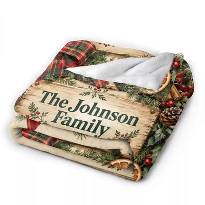 Folded personalized Christmas flannel blanket showing the rustic family name sign, festive plaid artwork, printed greenery, and white reverse side.