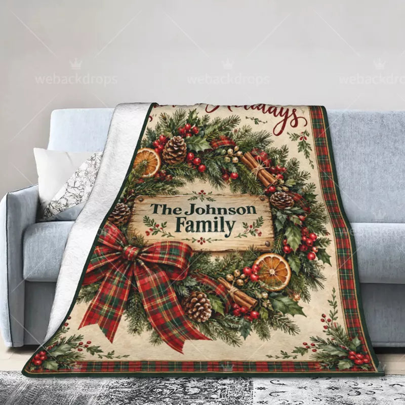 Personalized Christmas wreath blanket draped over a light gray sofa, displaying a plaid bow, evergreen branches, custom family name, and white reverse side.