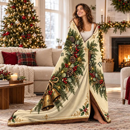 Woman wrapped in a personalized cedar wreath Christmas blanket with vintage bell artwork beside a decorated Christmas tree and fireplace.