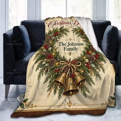 Personalized family Christmas blanket displayed on a dark blue sofa with an evergreen wreath, decorative brass-colored bells, and family name.