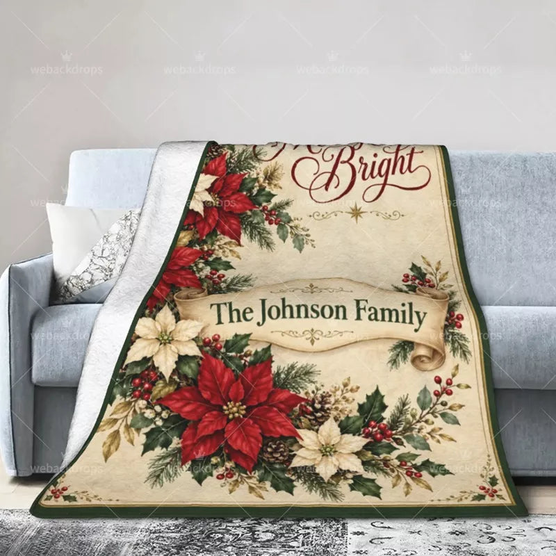 Personalized Christmas throw blanket draped over a gray sofa, displaying red and cream poinsettias, a family name ribbon, and a green border.