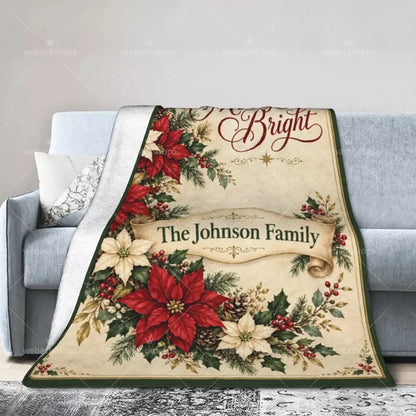 Personalized Christmas throw blanket draped over a gray sofa, displaying red and cream poinsettias, a family name ribbon, and a green border.
