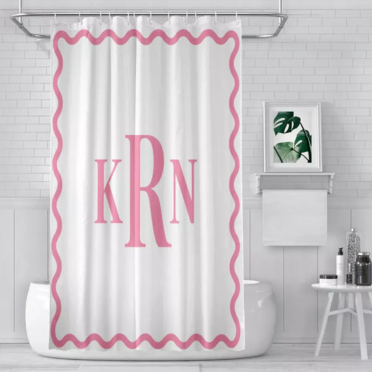 White shower curtain with pink wavy border and large KRN monogram hanging over a bathtub in a bright white tiled bathroom.